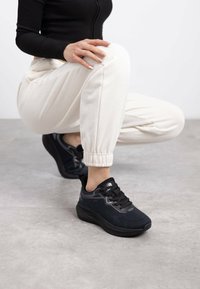 Las zapatillas deportivas negras presentan un upper de malla texturizada y una suela acolchada. Al combinarse con joggers blancos, las zapatillas cuentan con cordones y un branding sutil.
