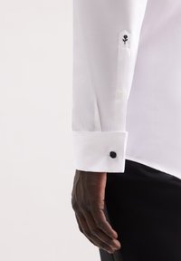 Camicia bianca con polsini a punta, dotata di un bottone nero e un piccolo design ricamato di una rosa nera. Tessuto liscio e taglio sartoriale.