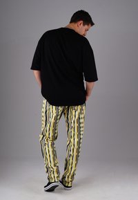 Man wearing black oversized t-shirt, patterned yellow and black pants, and black slip-on shoes, standing against a plain gray background.