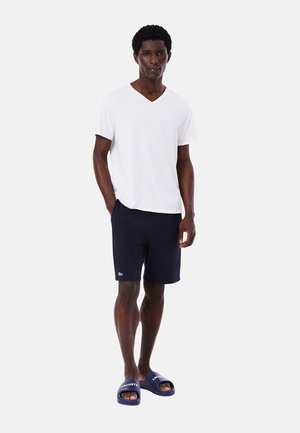 Camiseta blanca de cuello en V, pantalones cortos azul marino con bolsillos laterales, chanclas azules con logo blanco. Diseño simple, casual y ajustado.