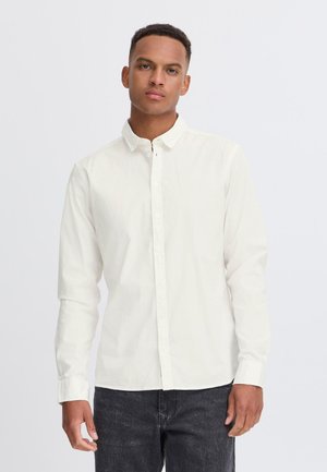 Ung mann iført hvit langermet knapp-skjorte og svarte jeans, stående mot en ensfarget lys bakgrunn.