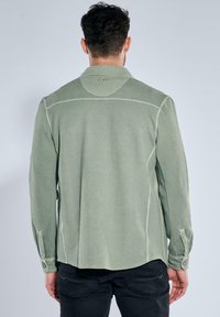 Green textured shirt with long sleeves, button cuffs, and a pointed collar, featuring contrast stitching and a high-low hem.