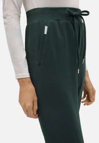 Pantalones de textura verde oscura con una cinturilla alta, lazo ajustable y bolsillos laterales con detalles en tono plateado.