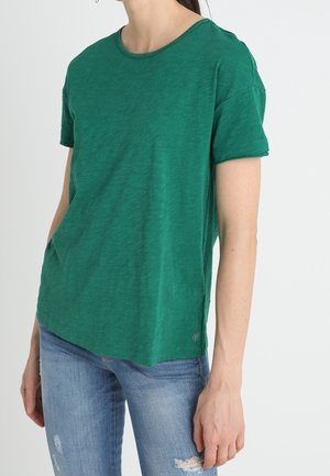 Osoba ubrana w gładki zielony t-shirt z krótkim rękawem i niebieskie podarte jeansy, stojąca z jedną ręką w kieszeni na białym tle.