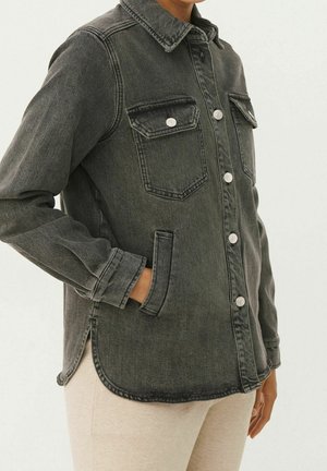 Person trägt eine ausgewaschene schwarze Jeansjacke mit Knöpfen, zwei Brustklappentaschen und eine Hand in einer seitlichen Schlitztasche.