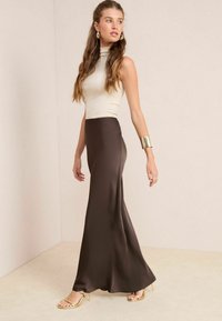 Een lange, satijnen bruine rok gecombineerd met een beige top met hoge nek. Kenmerkt zich door een gladde textuur en een relaxte pasvorm. Gouden ringoorbellen en sandalen accentueren de outfit.