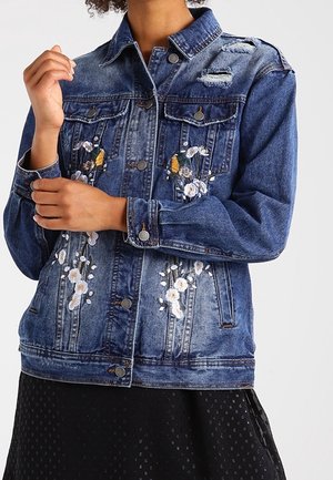 Jeansjacke mit abgenutzten Details und weißer und gelber Blumenstickerei, getragen über einem schwarzen, strukturierten Rock.