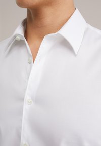 Bílá košile na knoflíky s klasickým límcem, vyrobená z hladké látky s jemnou strukturou. Knoflíky jsou světle zbarvené a mírně lesklé.