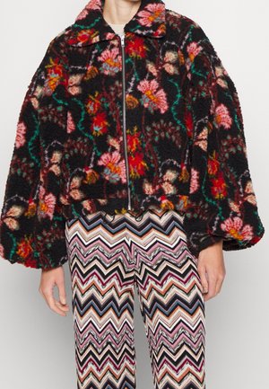 Chaqueta negra con cremallera con estampado floral colorido y mangas anchas, combinada con pantalones con patrón de zigzag en varios colores.