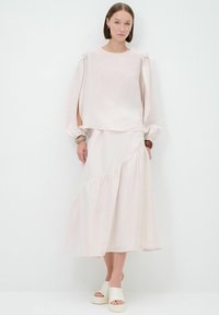 Blusa fluida rosa chiaro con maniche a palloncino e aperture, abbinata a una gonna a strati coordinata, entrambe realizzate in tessuto morbido e leggero. Sandali bianchi.