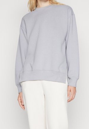 Kvinde iført en lysegrå langærmet sweatshirt og hvide bukser, stående mod en ensfarvet lys baggrund.