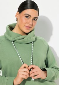 Felpa verde con collo avvolgente, dotata di un cordoncino bianco intrecciato. Tessuto morbido, vestibilità comoda e dettagli delle cuciture ben visibili.