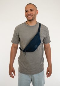 Navyblauer Stoff-Slingbag mit geschwungenem Design, Reißverschluss und verstellbarem Tragegurt, getragen über einem grauen T-Shirt.