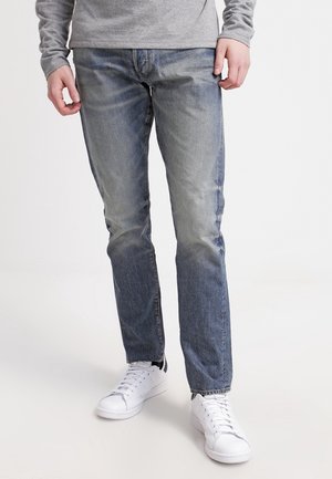 Mężczyzna w szarej koszuli z długim rękawem, wyblakłych niebieskich dżinsach i białych tenisówkach stoi na białym tle.