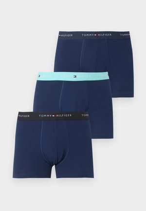 Atletický muž stojaci bosý, oblečený v tmavomodré boxerky Tommy Hilfiger na jednolivom bielom pozadí.