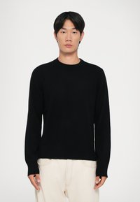 Sort sweater med lange ærmer og rund hals. Strikket tekstur og ribbet kant. Båret med lyse bukser.