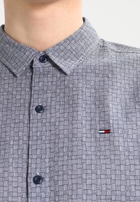 Camicia blu con motivo a quadri, colletto button-down e bottoni blu scuro. Presenta un piccolo logo rosso, bianco e blu vicino al petto.