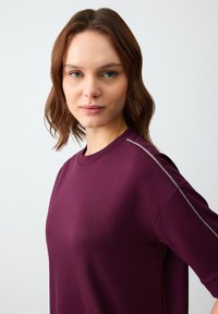 Bordeaux pullover met een ronde halslijn en korte mouwen. Heeft een zilveren accent op de schouder. Gladde textuur, relaxte pasvorm.