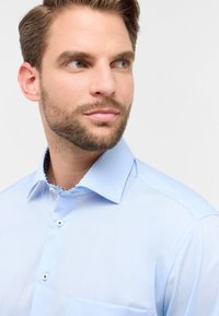 Camicia blu chiaro con colletto classico, pattina dei bottoni e taschino. Presenta un tessuto a fantasia contrastante all'interno del colletto.