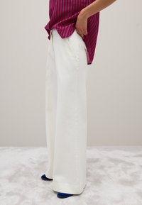 Persona che indossa pantaloni bianchi a gamba larga, scarpe blu e una maglietta larga a righe rosse e blu su uno sfondo chiaro e uniforme.