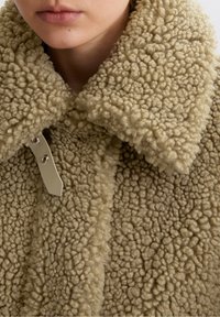 Beige fluffig kappa med en strukturerad, plyschig yta. Har en bred krage och silverdetaljer på stängningen.