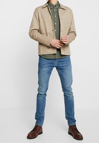 Beżowa bawełniana kurtka na zielonej koszuli zapinanej na guziki, w połączeniu z jasnoniebieskimi dżinsami i brązowymi skórzanymi butami. Prosty fason, zapięcie na guziki i kieszenie.