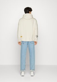 Roomkleurige oversized hoodie met een capuchon en trekkoord, geribbelde manchetten en geborduurde details, gecombineerd met lichtblauwe taps toelopende jeans en sneakers.