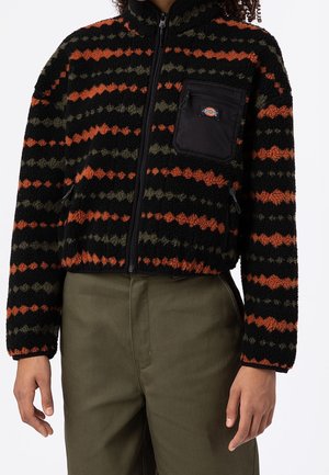 Chaqueta de forro polar negra con franjas horizontales en naranja y verde oliva, cremallera frontal, bolsillo negro en el pecho con logo, usada con pantalones verde oliva.
