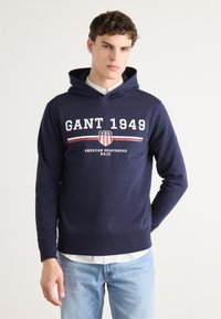 Tengerészkék kapucnis pulóver pamutból, fehér "GANT 1949" logóval, piros és fehér csíkokkal, valamint kenguruzsebbel. Fehér galléros ing felett viselve.