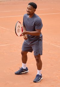Grå atletisk skjorte og shorts, hvide sokker og blå tennissko. Holder en rød tennisketsjer, står på en grusbane.