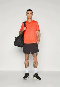 Mann iført knalloransje T-skjorte og svarte shorts, holder en svart treningsbag over skulderen, står mot en ensfarget bakgrunn.