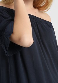 Marinblus med avskuret axel, tillverkad i löst, flödande tyg och med spetskantade ärmar. Mjuk textur och avslappnad passform som framhäver överarmen.