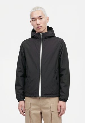 Un mannequin masculin porte une veste noire à capuche et à fermeture éclair avec un pantalon cargo beige, sur un fond blanc uni.