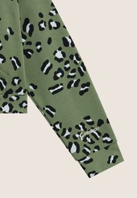Manche de sweat-shirt vert arborant un imprimé léopard en noir et blanc, avec un poignet côtelé et un logo brodé près de l'ourlet.