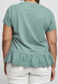 Ljusgrön bomullströja med korta ärmar, med en volangkant nertill. Baksidan har en slät textur och rundad halsringning. Tillsammans med vita byxor.