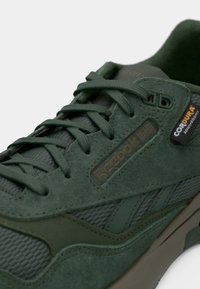 Detalhe de um ténis Reebok verde com texturas em camurça e malha, atacadores verdes e uma etiqueta de tecido Cordura na lateral.
