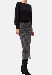 Schwarzes Langarmoberteil kombiniert mit einem enger geschnittenen, gemusterten Midirock in dunklen Farbtönen. Der Rock hat ein strukturiertes Design und wird mit knielangen schwarzen Stiefeln getragen.