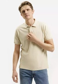 Beżowa koszulka polo wykonana z bawełny, o miękkiej teksturze, z kołnierzykiem, trzema guzikami i krótkimi rękawami. Noszona z niebieskimi dżinsami.