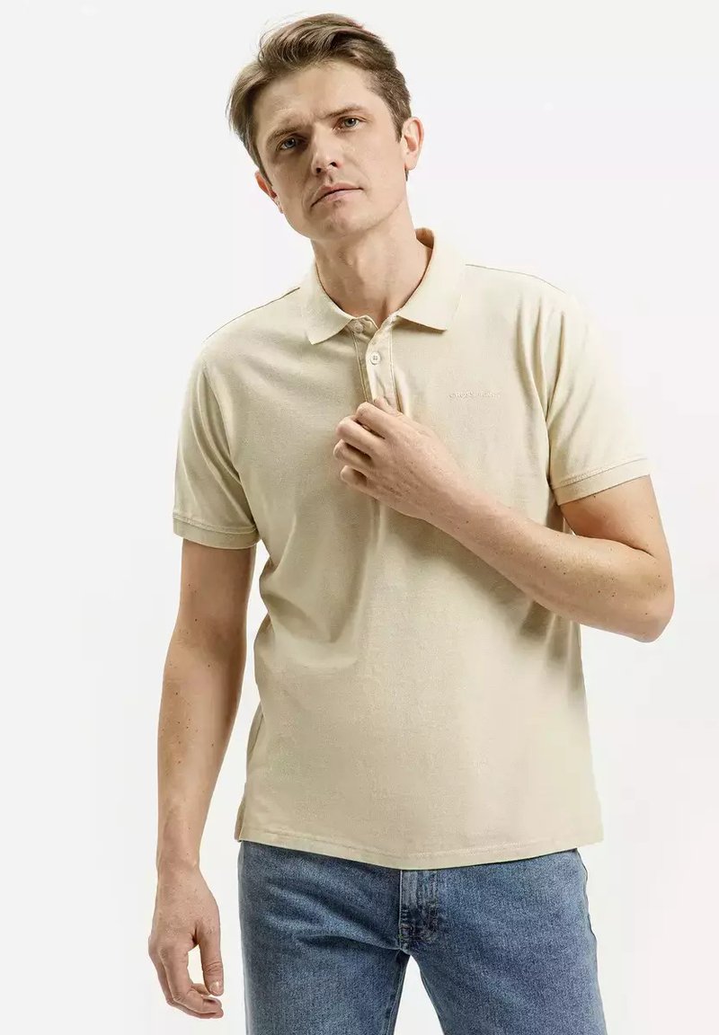 Beżowa koszulka polo wykonana z bawełny, o miękkiej teksturze, z kołnierzykiem, trzema guzikami i krótkimi rękawami. Noszona z niebieskimi dżinsami.