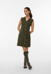 Olijfgroene corduroy jurk met een V-hals, mouwloos ontwerp, sluiting met knopen aan de voorkant en textuurstof, gecombineerd met kniehoge bruine laarzen.