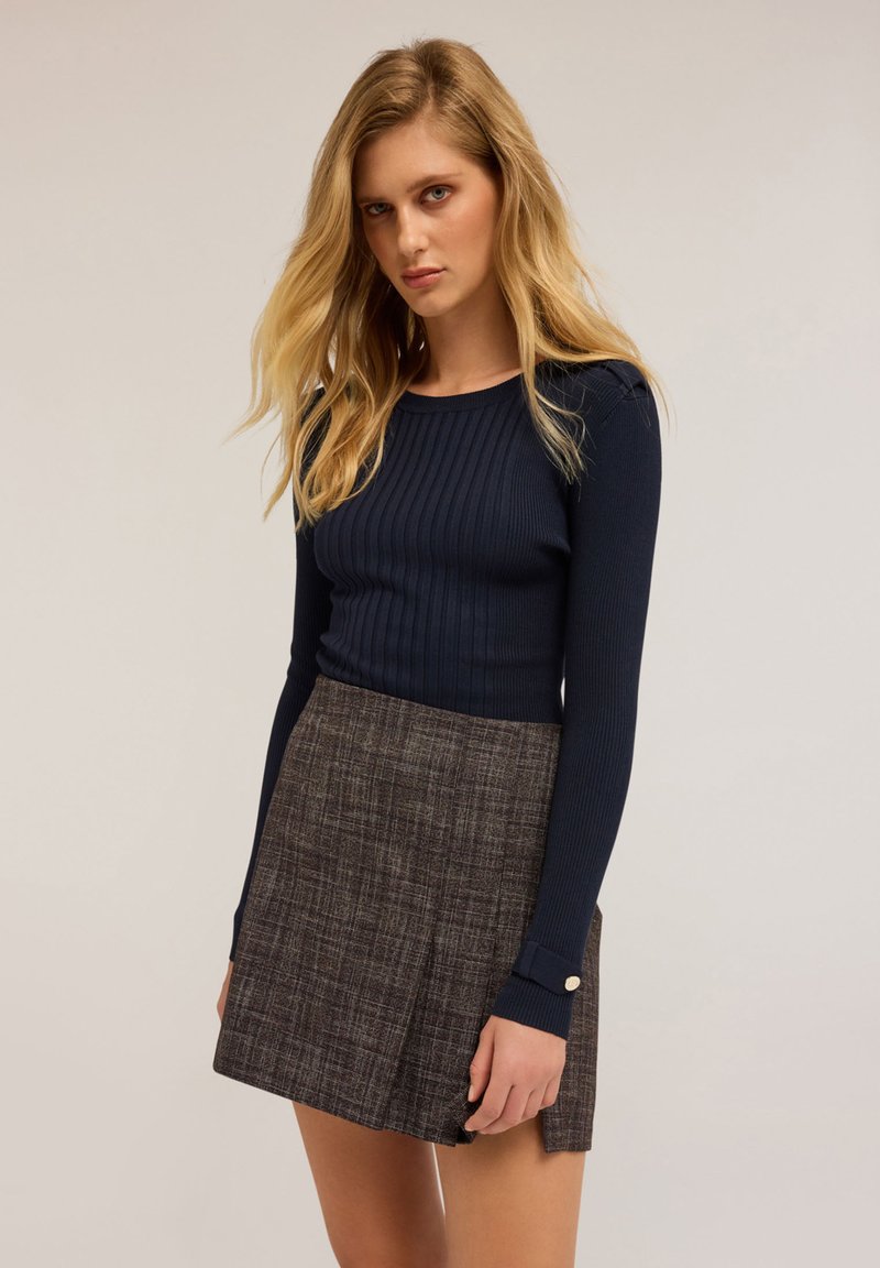 En blond kvinna i en marinblå randig långärmad topp och en mörk rutig minikjol framför en enfärgad bakgrund.