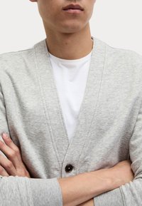 Cardigan grigio chiaro realizzato in un tessuto morbido, con scollatura a V, chiusura frontale con bottoni e maniche lunghe. T-shirt bianca sotto.