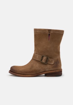 Botte en daim marron mi-mollet avec une sangle à boucle latérale et un petit talon en bois sur fond blanc uni.