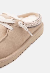 Beiger Wildleder-Slipper mit weißen Schnürsenkeln, gestricktem Besatz, Fleecefutter und Gummisohle, dargestellt aus einer nahen seitlichen Perspektive.