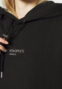 Hoodie noir avec une finition texturée, arborant un logo en filigrane argenté et un cordon de serrage avec des accents en métal argenté.