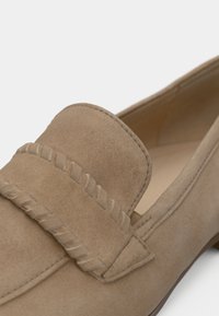 Καφέ δερμάτινο μοκασίνι από σουέντ με ραμμένη διακοσμητική ταινία και απαλή υφή. Η στρογγυλεμένη μύτη και η χαμηλή σόλα ενισχύουν το κλασικό σχέδιο.