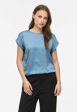 Frau mit langen braunen Haaren, die eine kurzärmelige blaue Satinbluse und schwarze Hose trägt, vor weißem Hintergrund stehend.