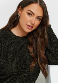 Pull en tricot vert foncé avec une texture côtelée et un col rond. Modèle avec de longs cheveux bruns, portant des boucles d'oreilles en argent.