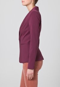 Femme debout de profil portant un blazer prune ajusté et un pantalon marron clair sur un fond gris uni.