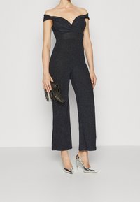 Musta, olkapäiltä avattu jumpsuit, jossa on tiukka vartalo ja leveät lahkeet. Kangas antaa hienovaraista kimallusta. Yhdistetty metallinvärisiin korokepolkimiin ja clutch-laukkuun.